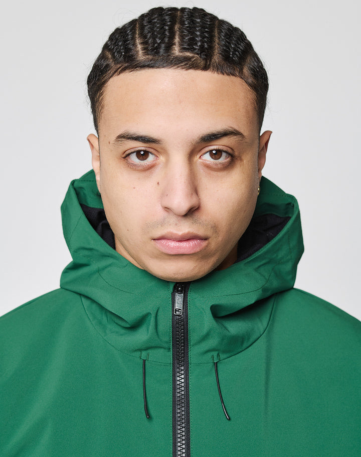 A person with braided hair wears the LIVE FAST DIE YOUNG Basic Shell Jacket with a water-repellent zipper and oversize fit, hood up, looking at the camera against a plain light background.