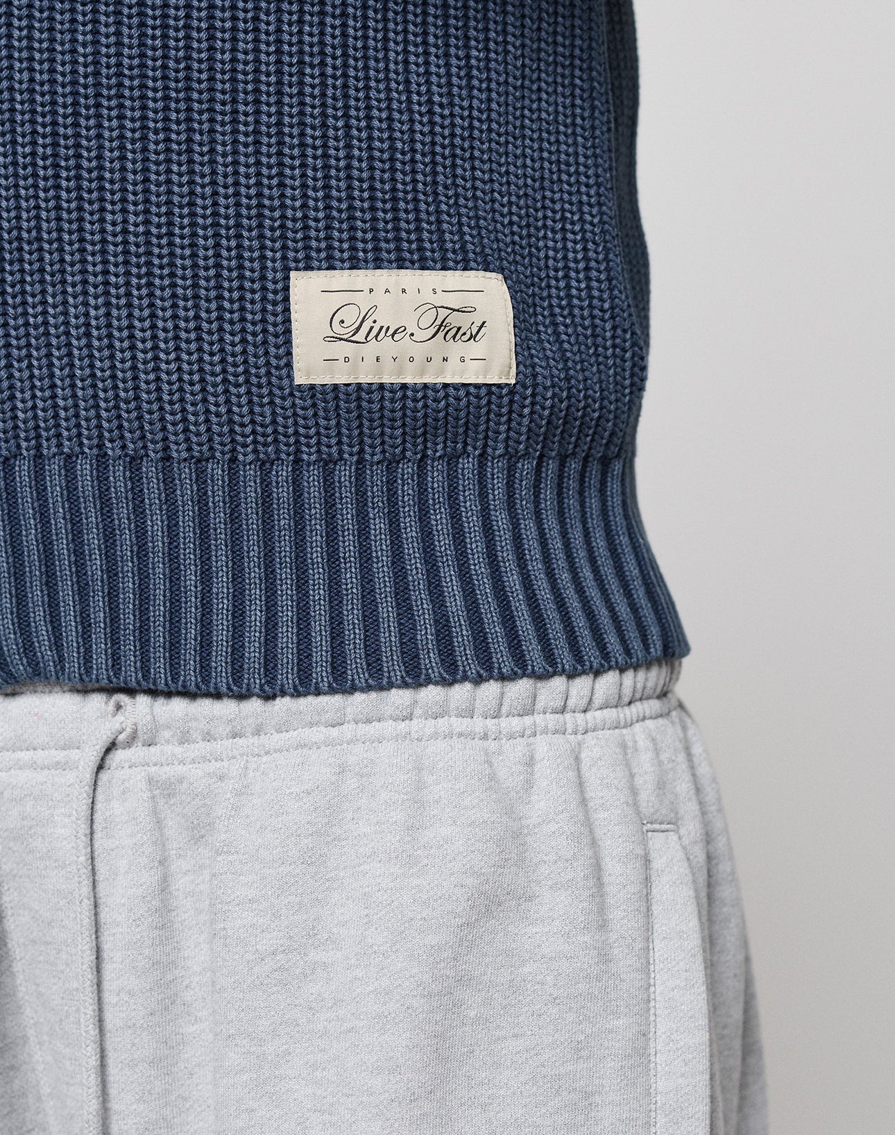 Close-up of a navy blue LFDY Knit Jacket by LIVE FAST DIE YOUNG with a Stehkragen and sewn-on Live Fast label, shown above light gray sweatpants with visible waistband and pocket.
