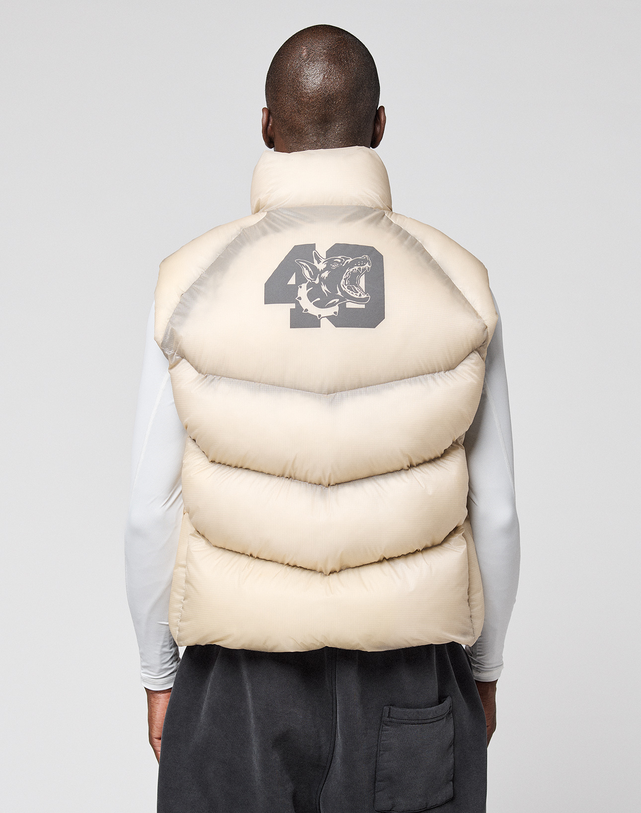 A person in the LIVE FAST DIE YOUNG Live Fast Puffer Vest—an oversized beige vest with a bold 40 and tiger graphic on the back—layered over a nylon jacket, stands facing away from the camera against a plain background.
