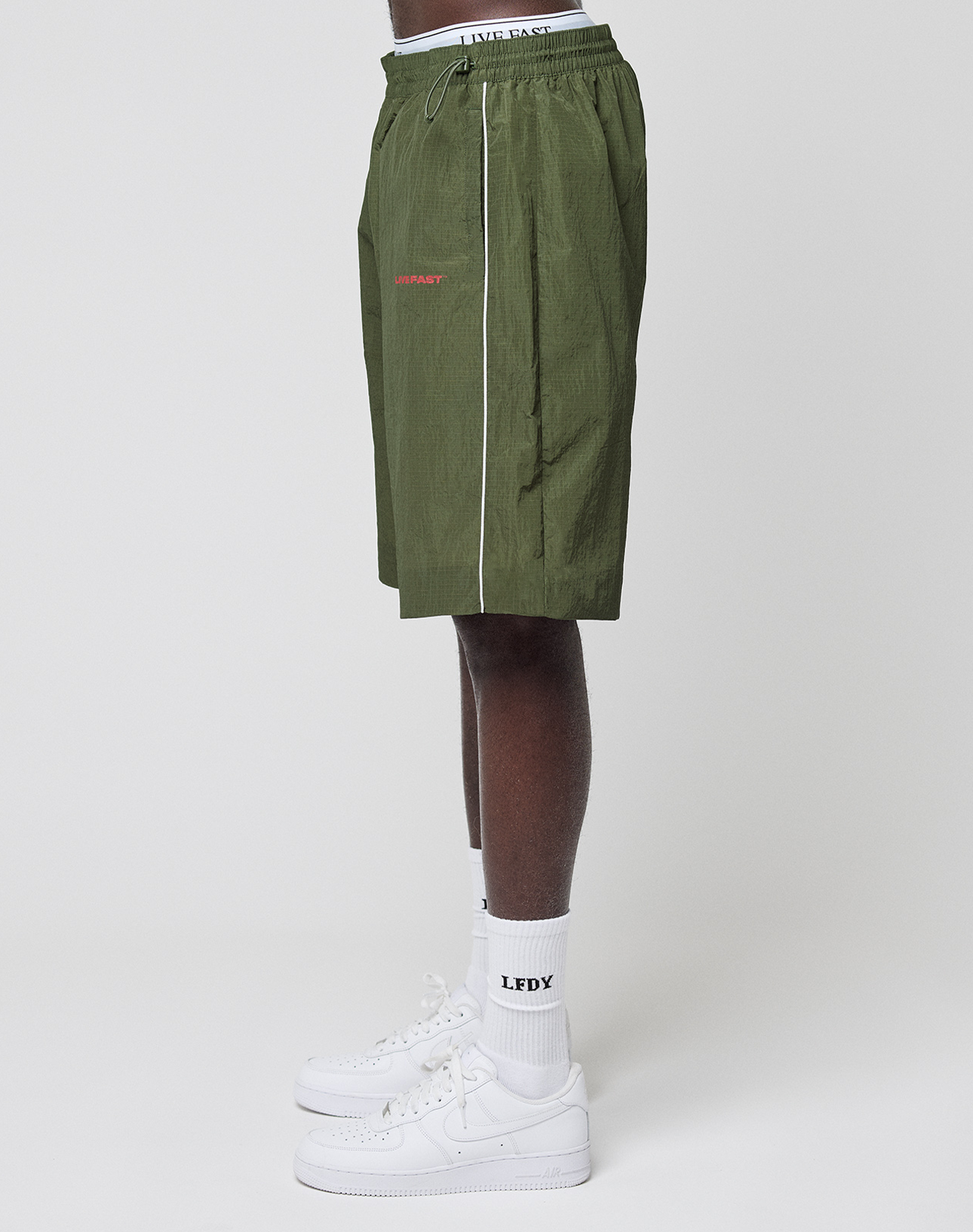 A person stands sideways in LFDY Nylon Trackshorts by LIVE FAST DIE YOUNG—olive green with a white stripe and "LIVE FAST" waistband—paired with LEDY white socks and white sneakers, against a plain light gray background.