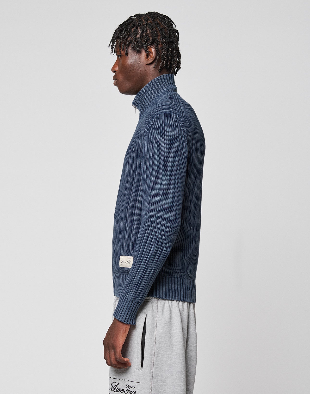 A person with short, twisted hair stands sideways against a plain white background, wearing the blue LFDY Knit Jacket by LIVE FAST DIE YOUNG and light gray sweatpants.