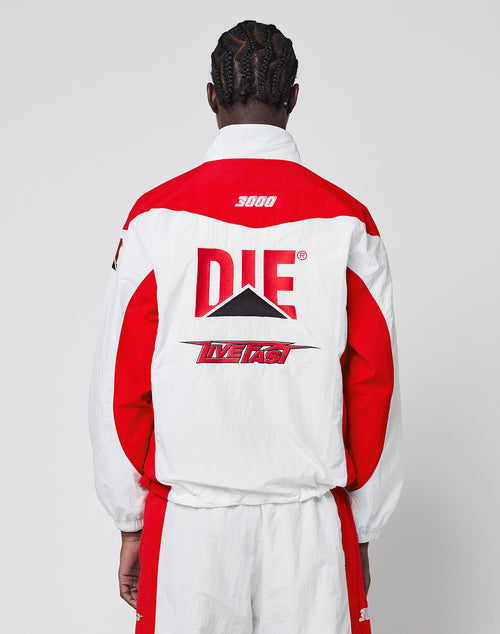 A person with braided hair, seen from behind, wears the LIVE FAST DIE YOUNG LFDY Trackjacket 3000—a regular fit red and white jacket with bold “DIE,” “LIVE FAST,” graphic designs, multiple logos, and the number 3000 near the collar.