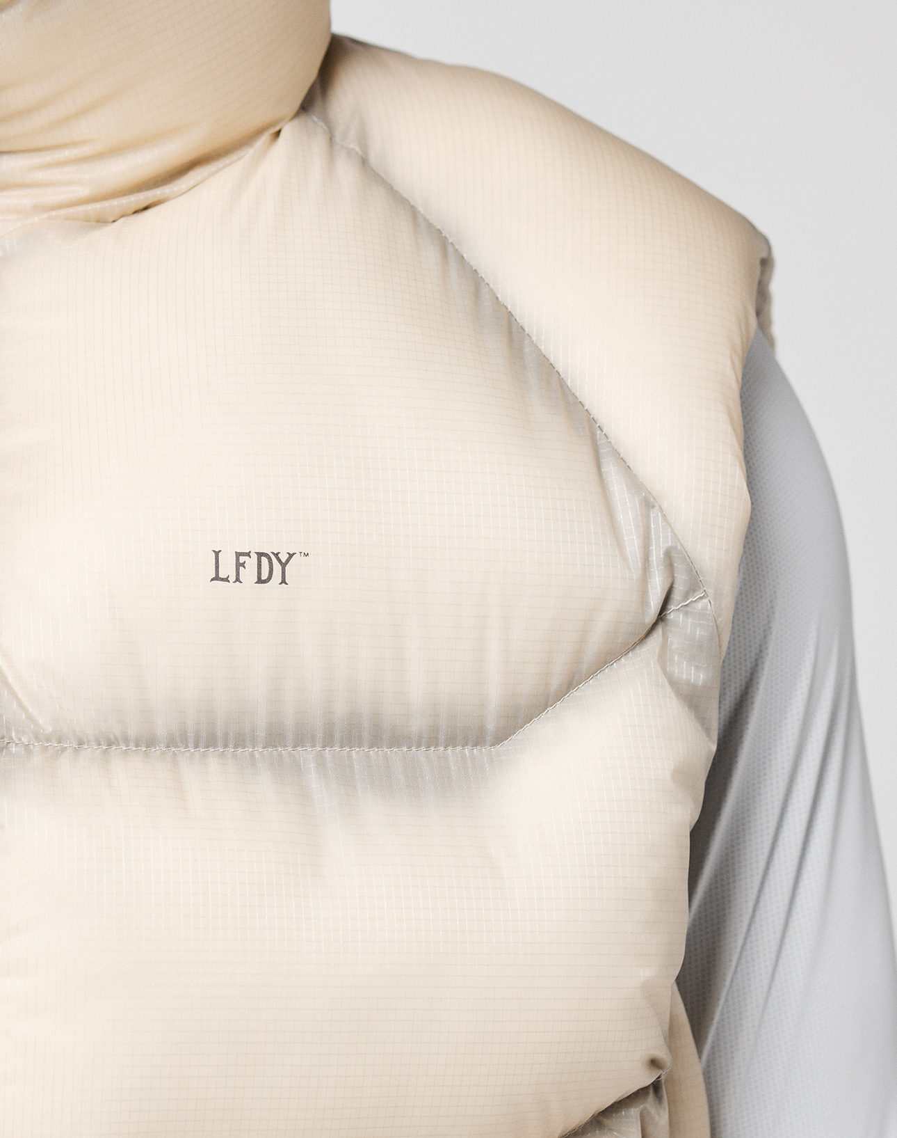 Close-up of a person wearing the LIVE FAST DIE YOUNG Live Fast Puffer Vest in light beige with transparent outer fabric and LFDY print, layered over a light gray long-sleeve shirt. Only the upper torso and part of the right arm in oversize fit are shown.