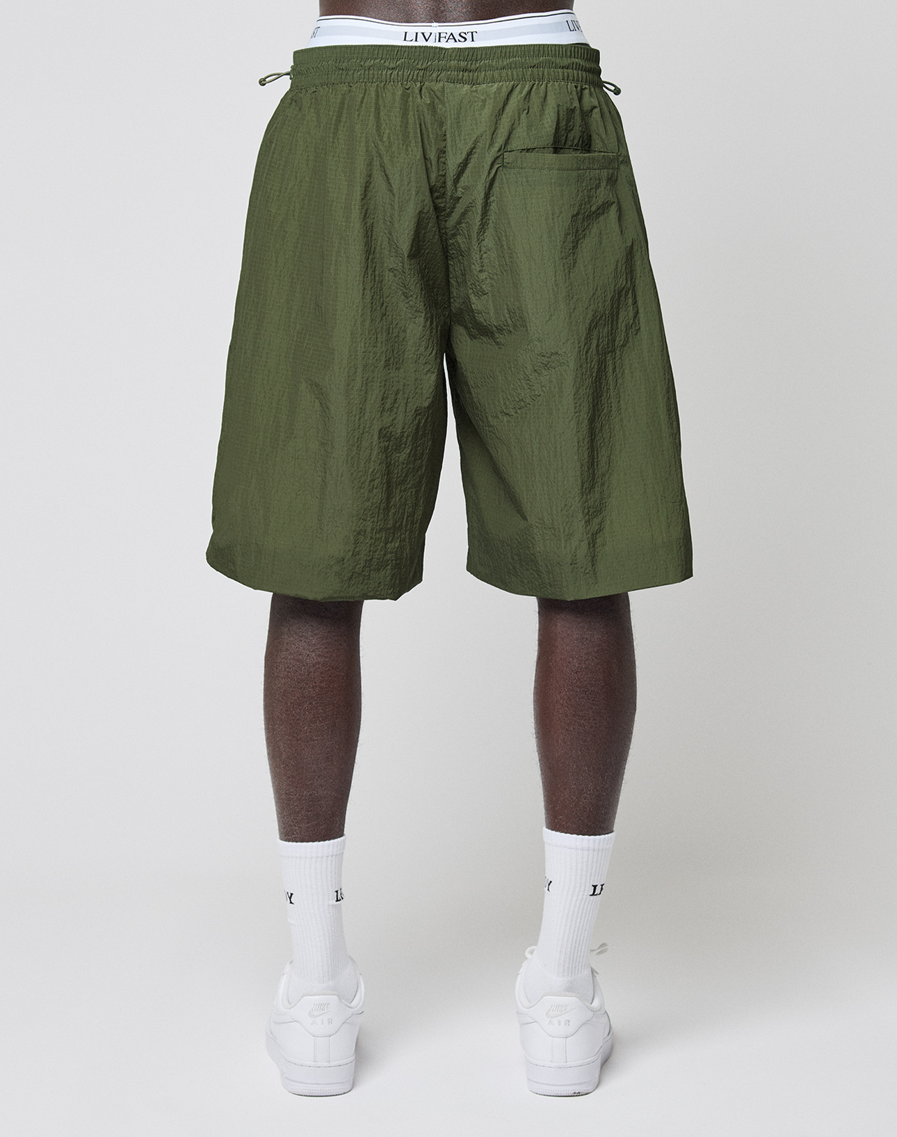 A person stands facing away, wearing LIVE FAST DIE YOUNG LFDY Nylon Trackshorts in olive green, white crew socks, and white sneakers. The shorts feature a back pocket with the logo-print waistband of their white underwear visible above.