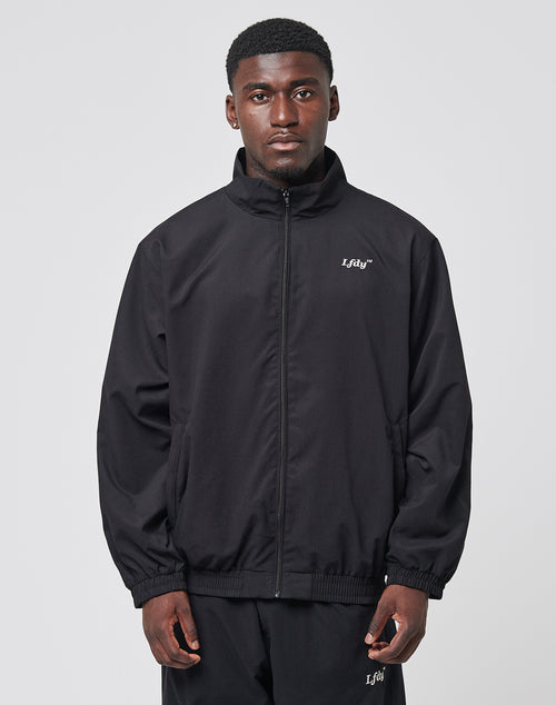 A man faces forward in an oversize black LFDY Trackjacket and matching pants by LIVE FAST DIE YOUNG, both featuring small white Lifay logos and subtle Logo-Stick details. The polyester outfit is set against a plain, light backdrop.