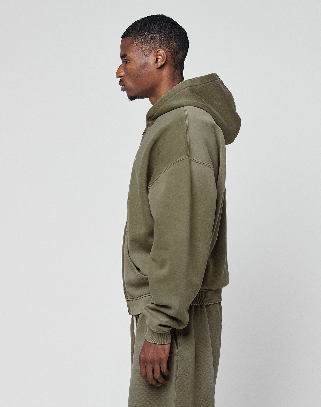 A man in profile facing left wears a LIVE FAST DIE YOUNG Basic Hooded Zip in olive green with a logo print and matching oversize sweatpants, set against a plain light gray background.
