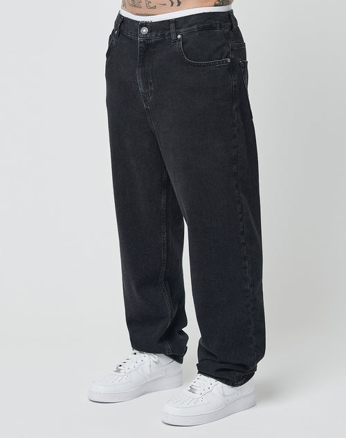 A person stands against a plain light background wearing LIVE FAST DIE YOUNG Wide Denim jeans in black with a relaxed, regular fit and classic five-pocket design, paired with white sneakers. Their torso is not fully visible.