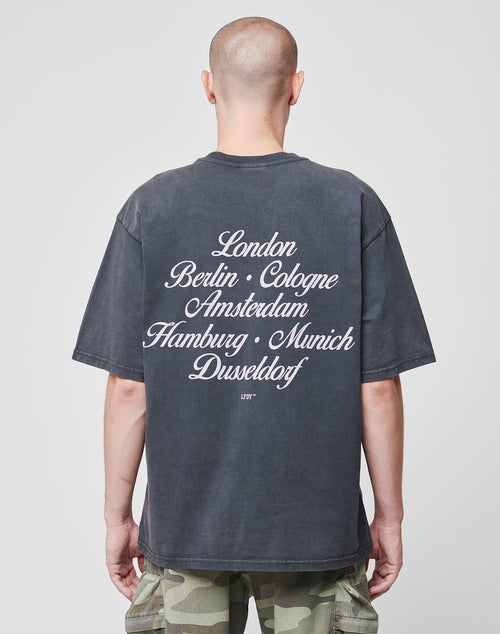 A person with a shaved head, wearing the LIVE FAST DIE YOUNG Black Chrome Tee with a city backprint—London, Berlin, Cologne, Amsterdam, Hamburg, Munich, Düsseldorf—faces away from the camera in camouflage pants.