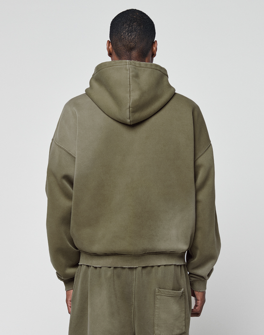 A person faces away from the camera in an olive green LIVE FAST DIE YOUNG Basic Hooded Zip and matching sweatpants. The hoodie is up, showing a subtle logo print, hands relaxed at their sides against a plain light backdrop.