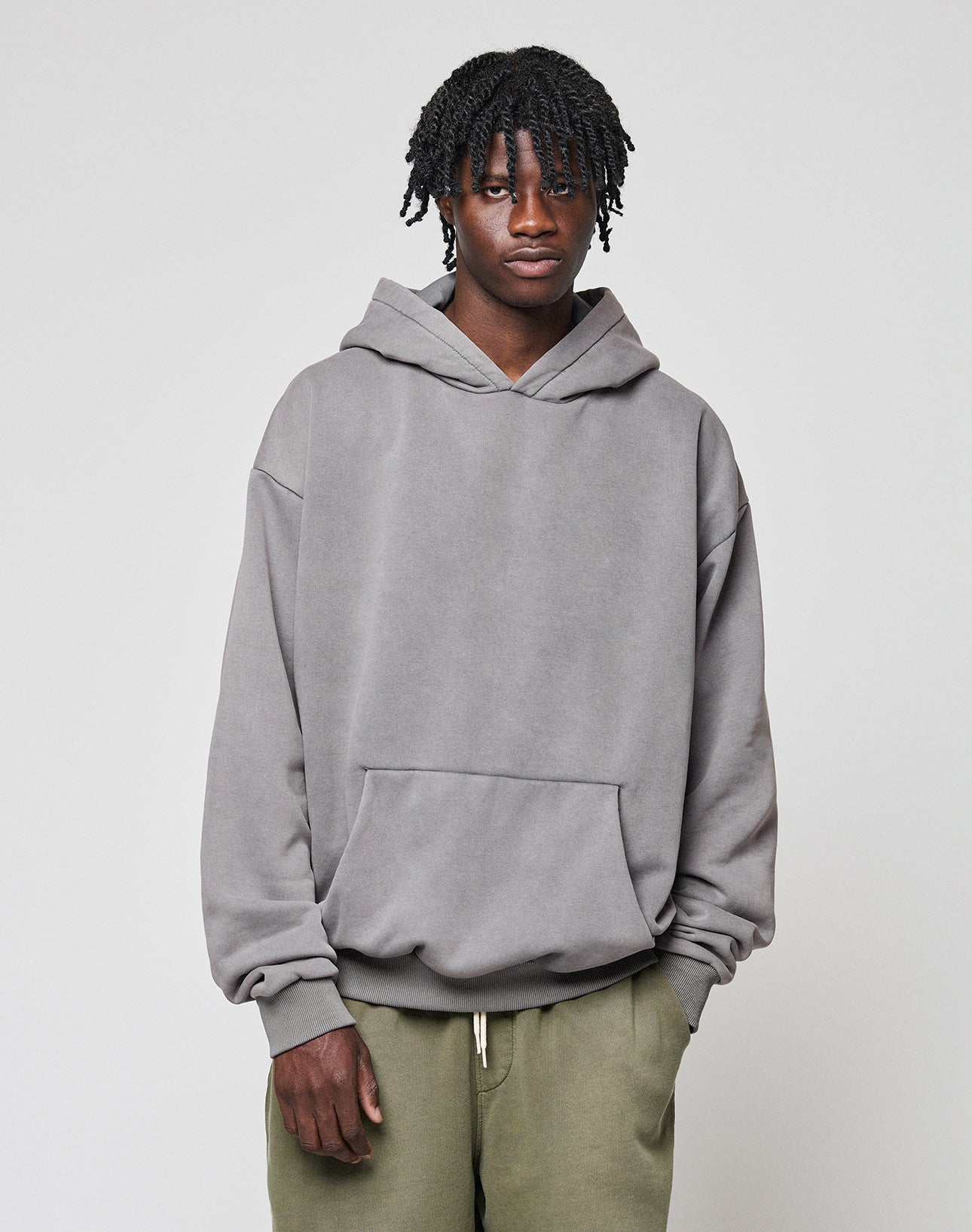 A person with short, twisted hair wears a loose-fitting gray LIVE FAST DIE YOUNG Basic Blank Hooded in Oversize Fit and olive green pants, standing against a plain light gray background.