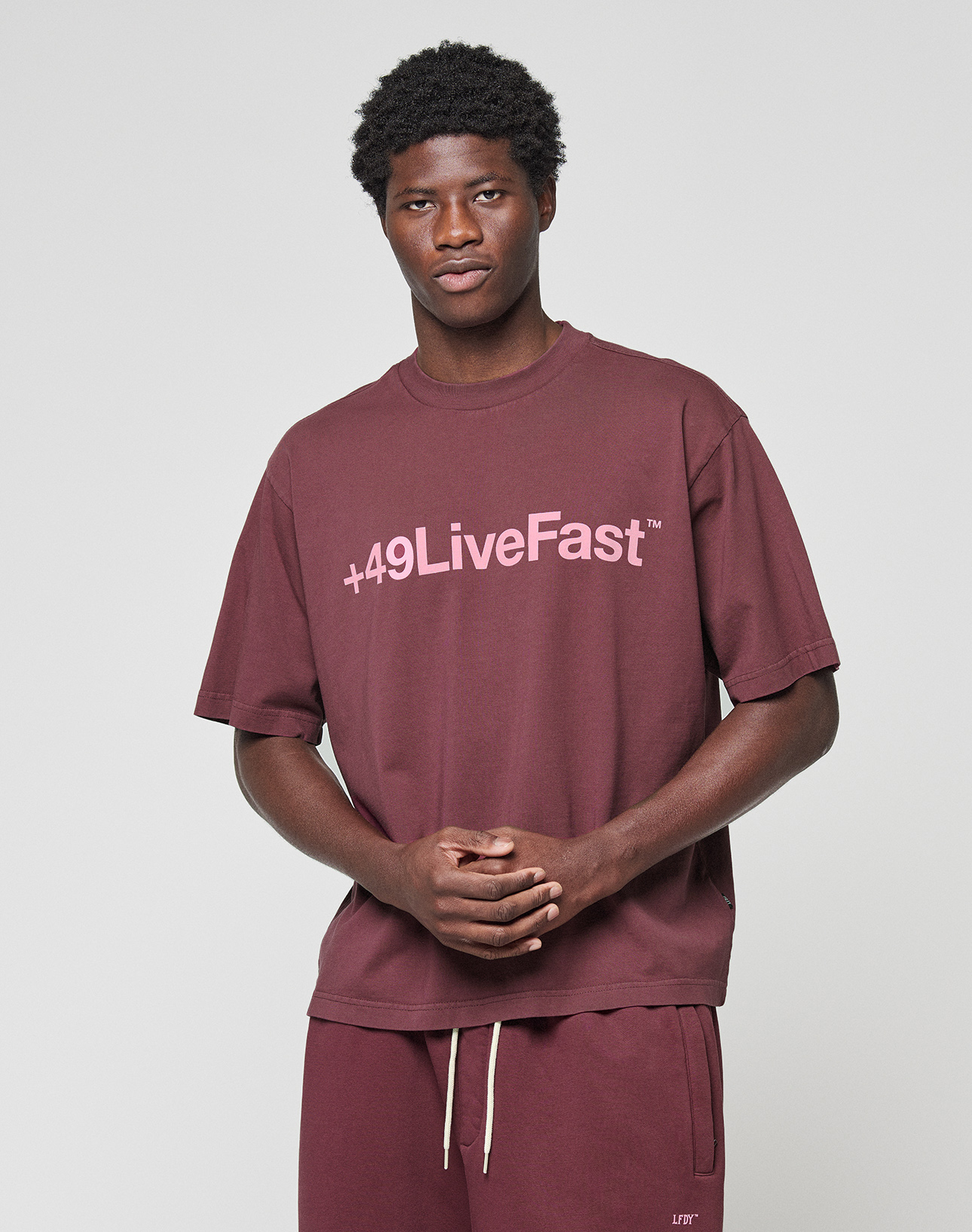 A man stands against a plain background wearing the LIVE FAST DIE YOUNG Basic +49 Tee in maroon with a light pink +49LiveFast™ front print, paired with shorts. He looks at the camera, hands clasped.