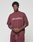 A man stands against a plain background wearing the LIVE FAST DIE YOUNG Basic +49 Tee in maroon with a light pink +49LiveFast™ front print, paired with shorts. He looks at the camera, hands clasped.