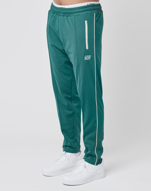 A person in teal green LIVE FAST DIE YOUNG Live Fast Trackjogger with a white zipper pocket, white piping, and subtle logo detail pairs it with white sneakers against a plain white background.