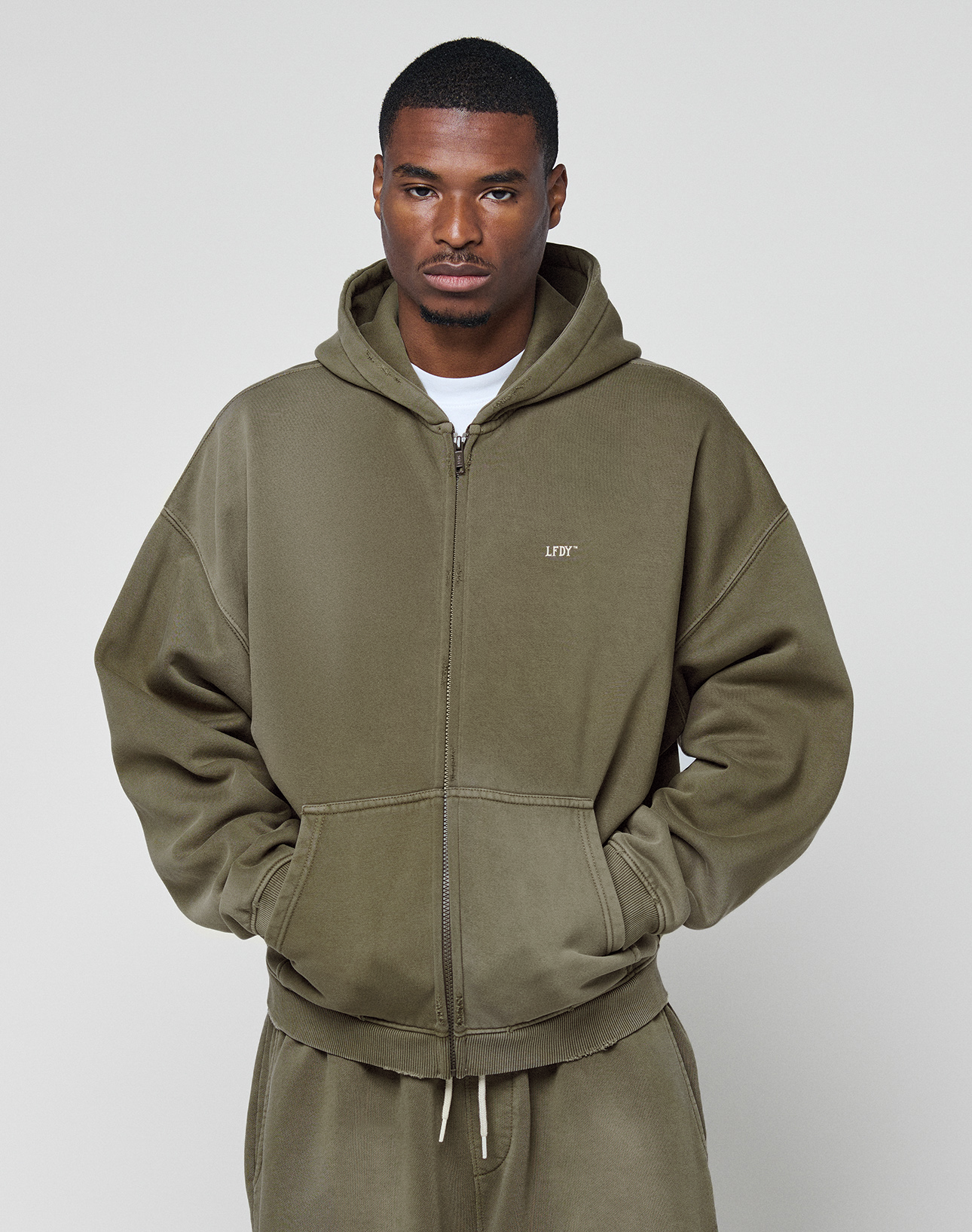 A man stands with his hands in his pockets, wearing the LIVE FAST DIE YOUNG Basic Hooded Zip and matching olive green sweatpants with an oversize fit, looking directly at the camera against a plain light background.