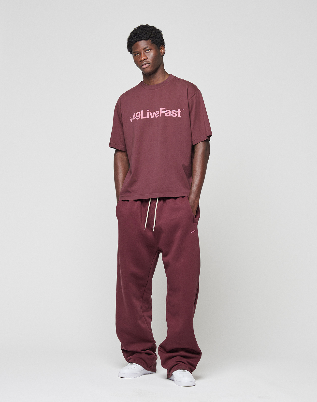 A man stands against a plain background, relaxed with hands by his sides, wearing the LIVE FAST DIE YOUNG Basic +49 Tee in an oversized maroon fit with frontprint and matching sweatpants. Made from 100% Baumwolle. White sneakers complete the look.