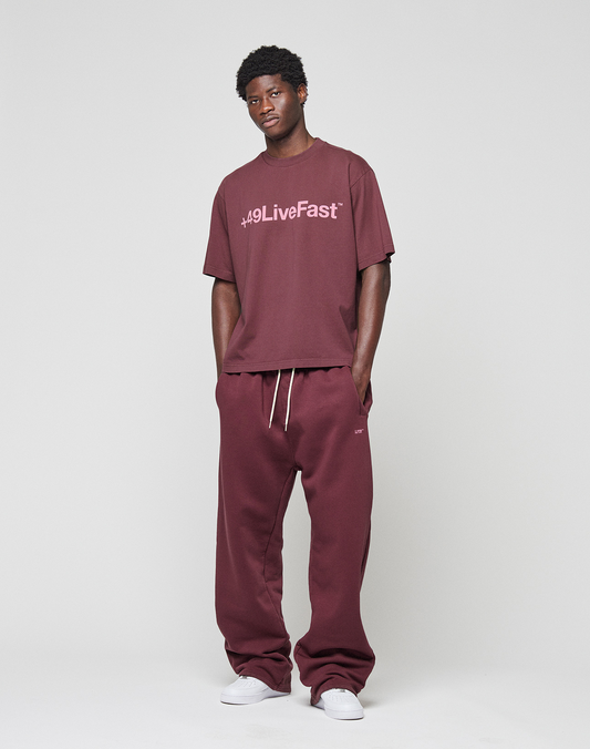 A man stands against a plain background, relaxed with hands by his sides, wearing the LIVE FAST DIE YOUNG Basic +49 Tee in an oversized maroon fit with frontprint and matching sweatpants. Made from 100% Baumwolle. White sneakers complete the look.