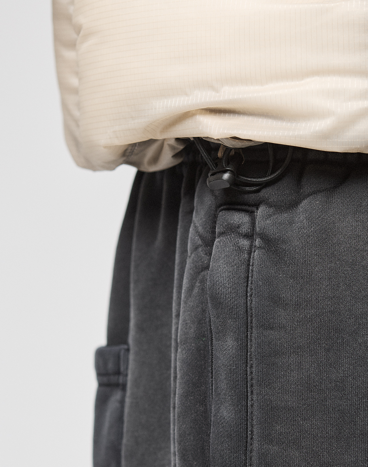 Close-up of a person wearing the Live Fast Puffer Vest by LIVE FAST DIE YOUNG, featuring a beige transparenter Oberstoff, paired with dark gray fleece sweatpants with an adjustable waistband drawstring.