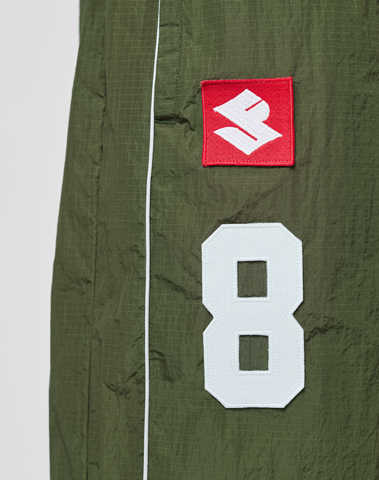 Close-up of LFDY Nylon Trackshorts by LIVE FAST DIE YOUNG, showing green polyamide fabric with a white number 8 patch and a red rectangular patch featuring a white stylized S, separated by a vertical white seam.