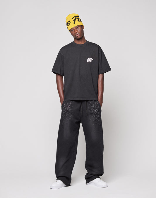 A man stands against a plain background wearing a loose, black LFDY Logo Tee by LIVE FAST DIE YOUNG, black speckled sweatpants, white sneakers, and a yellow beanie with black text.