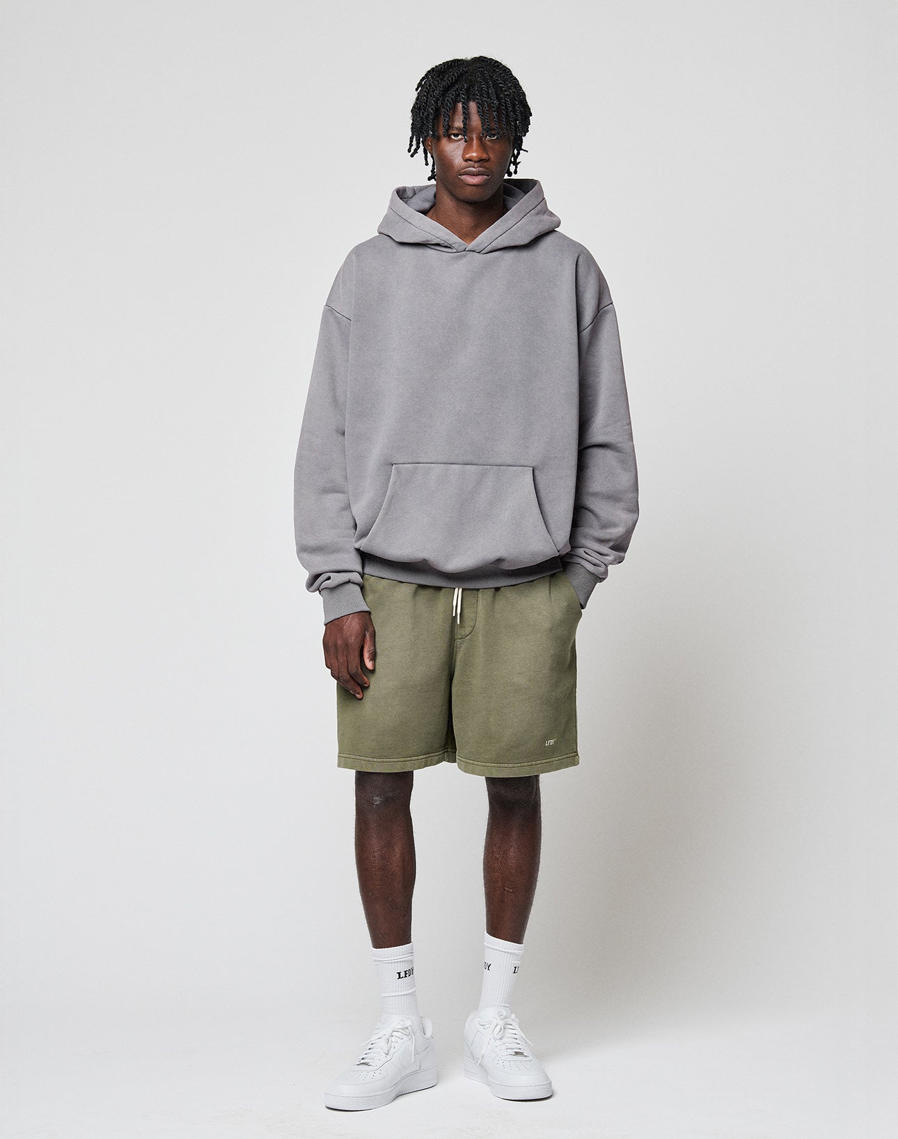 A man stands against a plain light background, wearing the LIVE FAST DIE YOUNG Basic Blank Hooded in Oversize Fit, olive green shorts, white crew socks, and white sneakers. He faces the camera with a neutral expression.