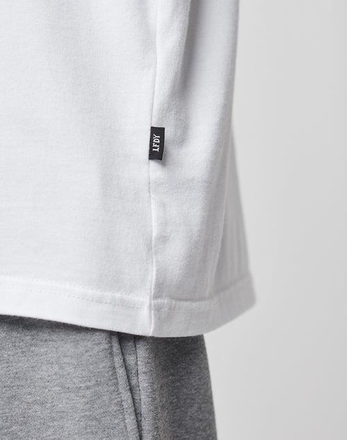 Close-up of a white LFDY Blank Tee by LIVE FAST DIE YOUNG, featuring a small black LFOT tag near the hem, styled with light gray sweatpants. The background is plain and light-colored.