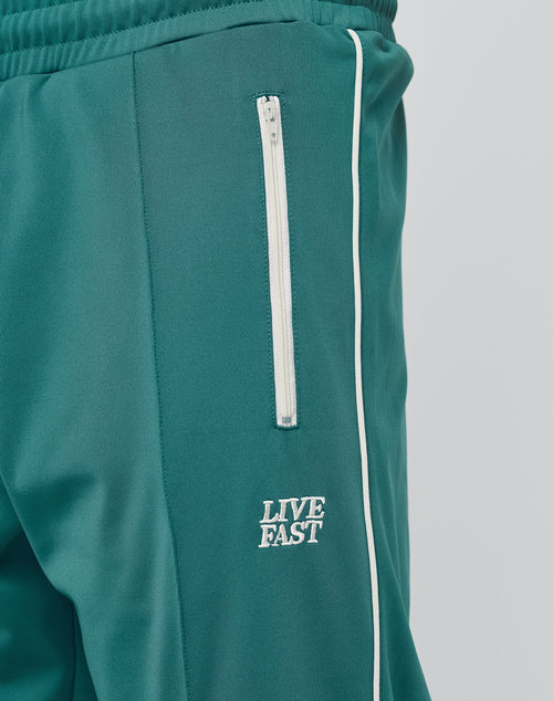 Close-up of the teal Live Fast Trackjogger by LIVE FAST DIE YOUNG, featuring a white zippered pocket, white piping, LIVE FAST embroidery on the upper thigh, and subtle Bügelfalte detailing for classic style.