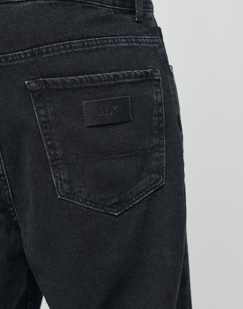 Close-up of the back pocket of black Wide Denim by LIVE FAST DIE YOUNG, featuring a black leather patch embossed with the brand name. The fabrics texture and detailed stitching are clearly visible.