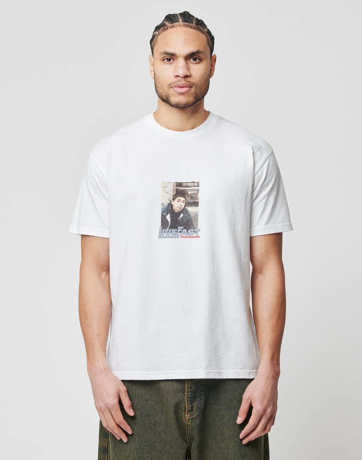 A man with braided hair wears the LIVE FAST DIE YOUNG Cope South Bronx Tee, a Regular Fit white T-shirt made from 100% Baumwolle, featuring a small rectangular graphic and Life Fast text on the front, standing against a light background.