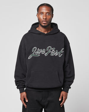 A man with braided hair faces forward in a plain light gray setting, wearing the LIVE FAST DIE YOUNG LFDY Script Hooded. The black 100% Baumwolle hoodie has Distress Effekte, an Oversize Fit, and Live Fast in script on the front.