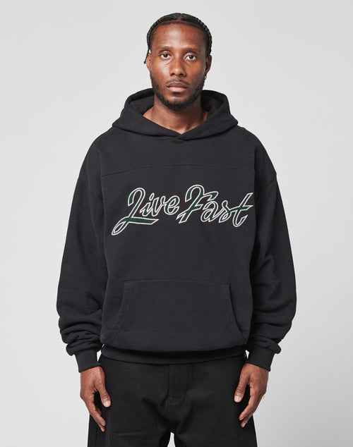 A man with braided hair faces forward in a plain light gray setting, wearing the LIVE FAST DIE YOUNG LFDY Script Hooded. The black 100% Baumwolle hoodie has Distress Effekte, an Oversize Fit, and Live Fast in script on the front.