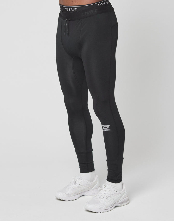 A person wearing Live Fast Performance Tights by LIVE FAST DIE YOUNG, in black slim fit with branding and elastic waistband, paired with white sneakers, stands against a plain white background. Their upper body is not visible.