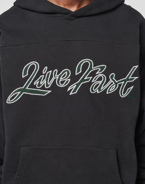 Someone wearing an oversize black LFDY Script Hooded hoodie by LIVE FAST DIE YOUNG, featuring Live Fast in large white and green cursive letters across the chest.