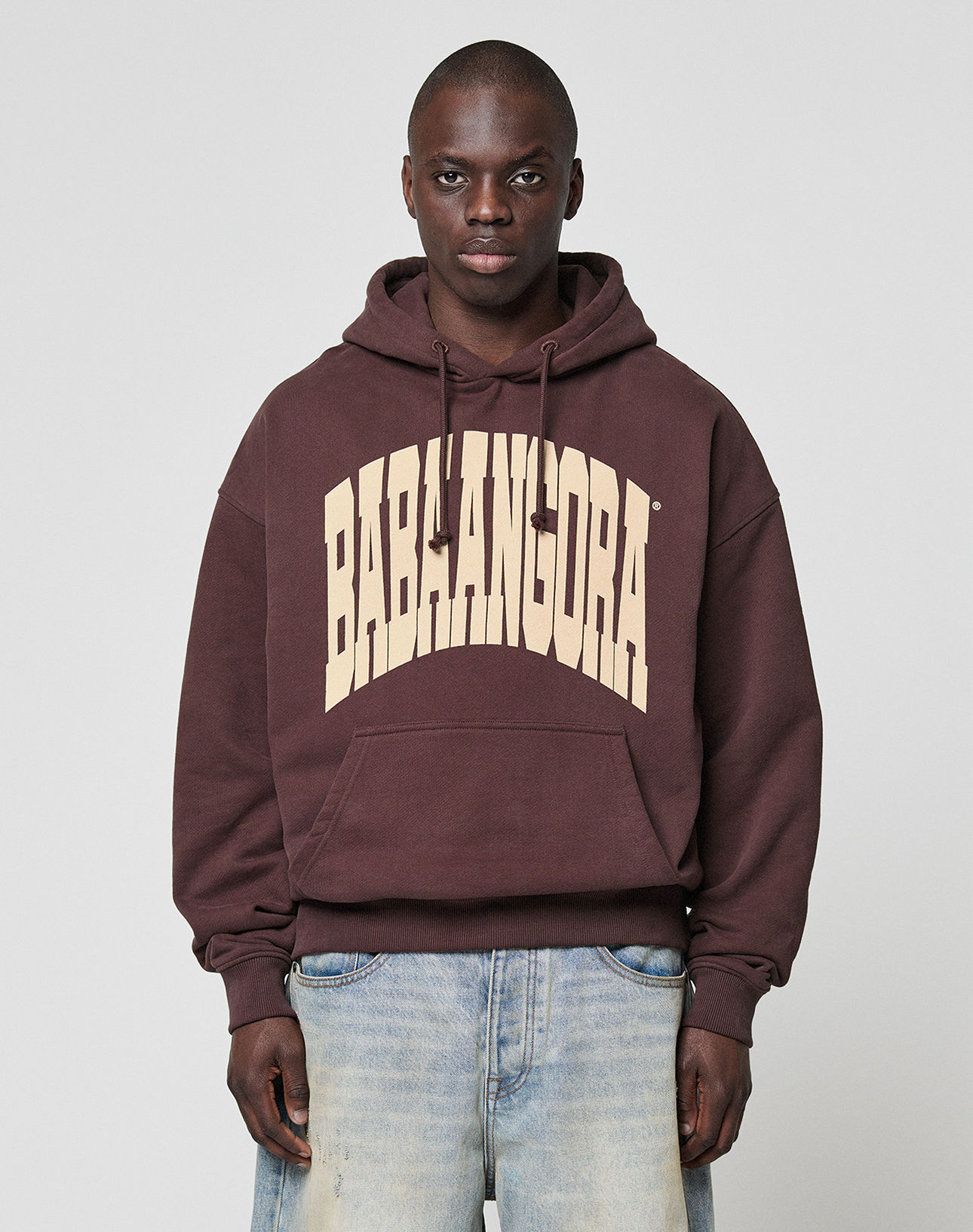 A person stands facing forward against a plain light gray background, wearing the LIVE FAST DIE YOUNG LFDY x Baba Angora Hoodie with large beige BABALANGORA letters on the chest, paired with light blue jeans.