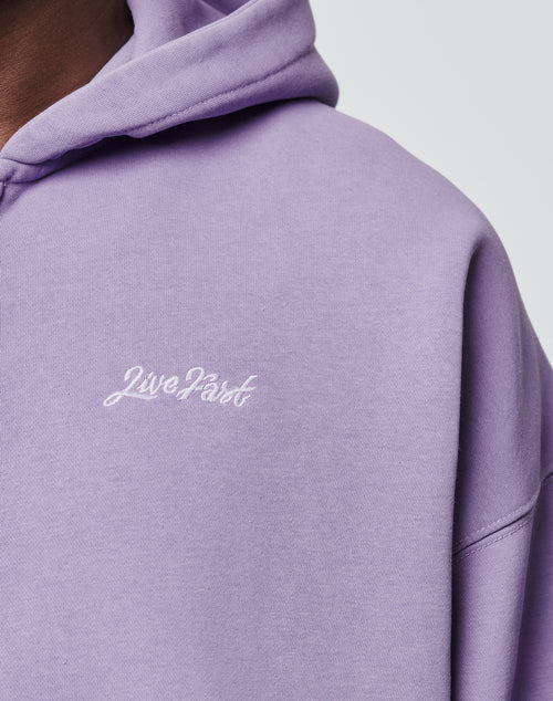 Close-up of a person wearing the LIVE FAST DIE YOUNG Basic Script Hooded Zip in light purple Vintage-Waschung, featuring Live Fast embroidered in white script on the chest; image shows neck, shoulder, and upper torso.