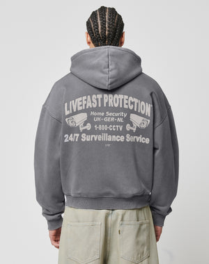 A person wearing the gray LFDY Protection Hooded Zip by LIVE FAST DIE YOUNG with front and back prints—LIVEFAST PROTECTION, Home Security UK-GER-NL, 1-800-CCTV, CCTV camera graphics. The cropped oversized fit has a hood with embroidered eyelets. Person faces away.