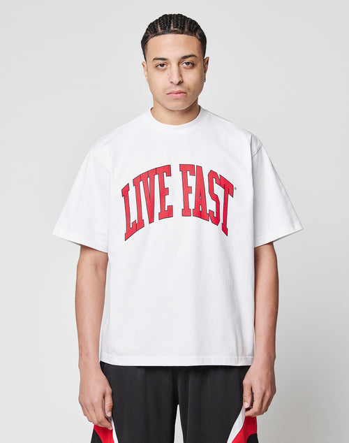 A person with braided hair wears the oversized Live Fast College Tee in loose white 100% Baumwolle, featuring a bold red LIVE FAST front print, paired with black and red shorts against a plain white background.