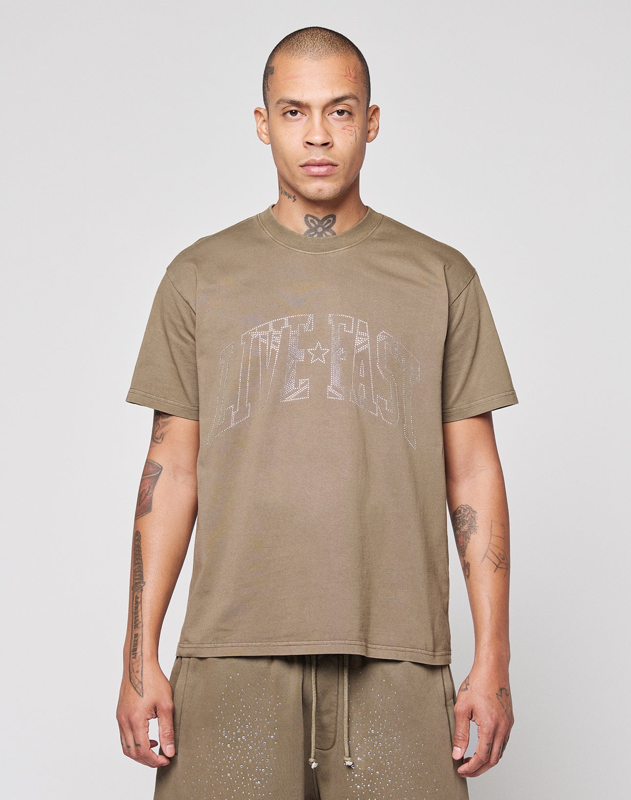 A person with closely shaved hair faces forward, wearing the LIVE FAST DIE YOUNG Live Fast Rhinestone Tee in oversize fit and matching brown shorts. Their tattooed arms are visible against a plain, light background.