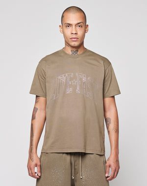 A person with closely shaved hair faces forward, wearing the LIVE FAST DIE YOUNG Live Fast Rhinestone Tee in oversize fit and matching brown shorts. Their tattooed arms are visible against a plain, light background.