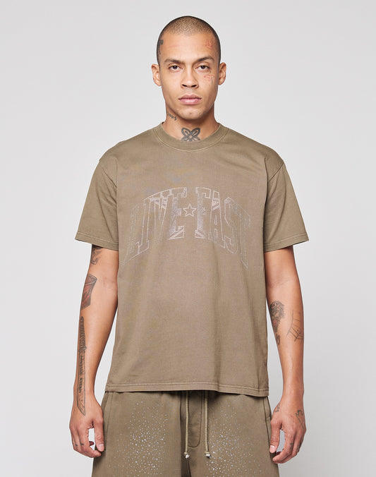 A person with closely shaved hair faces forward, wearing the LIVE FAST DIE YOUNG Live Fast Rhinestone Tee in oversize fit and matching brown shorts. Their tattooed arms are visible against a plain, light background.