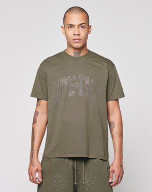 A tattooed person is wearing the LIVE FAST DIE YOUNG Live Fast Rhinestone Tee in olive green with matching shorts, standing against a plain light gray background.