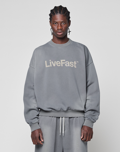 A person faces forward in a Basic LiveFast Sweater by LIVE FAST DIE YOUNG and matching gray sweatpants, both with a subtle faded look and the bold "LiveFast" logo, set against a plain light gray background.