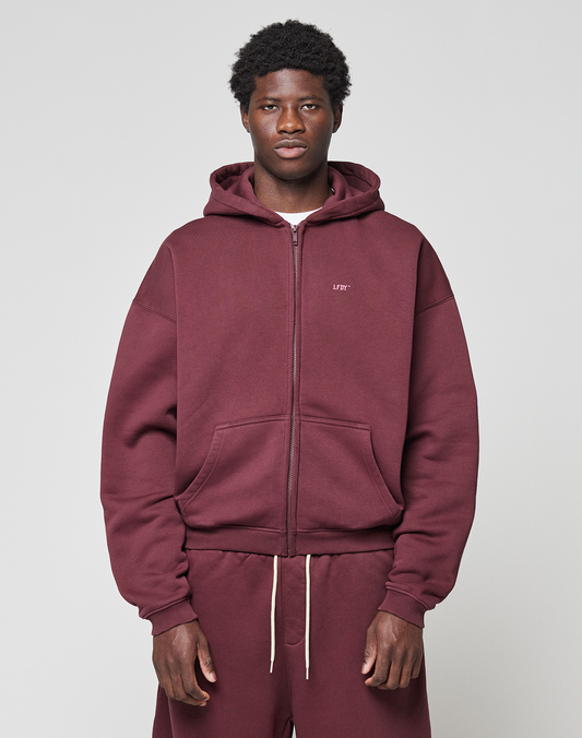 A man faces the camera in a maroon LIVE FAST DIE YOUNG Basic Hooded Zip with an oversized fit and matching sweatpants. Subtle branding details stand out against a plain, light gray background.