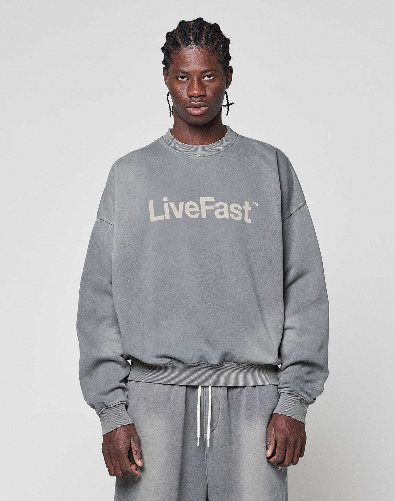 A man with braided hair wears the Basic LiveFast Sweater by LIVE FAST DIE YOUNG, an oversize fit gray sweatshirt with a handgefertigter Fade-Effekt and "LiveFast" print, paired with gray sweatpants against a light gray background.