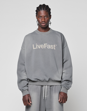A man with braided hair wears the Basic LiveFast Sweater by LIVE FAST DIE YOUNG, an oversize fit gray sweatshirt with a handgefertigter Fade-Effekt and "LiveFast" print, paired with gray sweatpants against a light gray background.