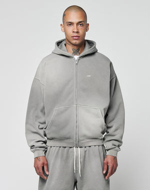 A person with a shaved head and neck tattoos models the LIVE FAST DIE YOUNG Basic Hooded Zip in gray, along with matching sweatpants, standing against a plain light background and looking at the camera.
