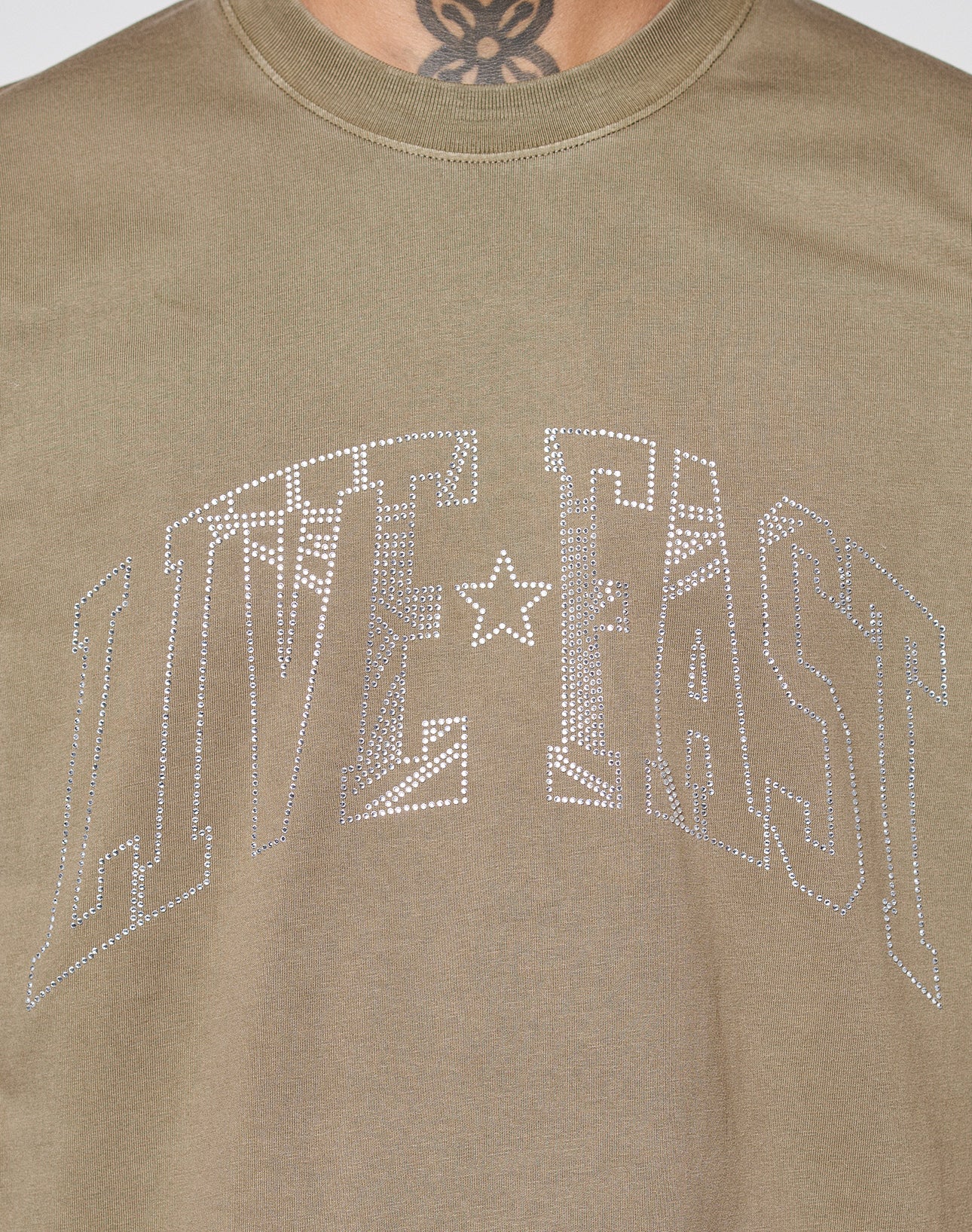 Close-up of the LIVE FAST DIE YOUNG Live Fast Rhinestone Tee in khaki vintage wash, featuring large rhinestone "LIVE FAST" lettering on the front. The model’s neck tattoo is partially visible above the oversized neckline.