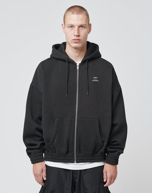 A young man with short blond hair wears the LF Vulgar Hooded Zip by LIVE FAST DIE YOUNG, an oversized black hoodie styled over a white t-shirt and black pants. He faces forward with a neutral expression against a plain light gray background.