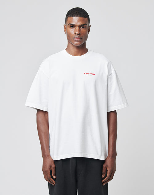 A person faces forward wearing the LFDY Cinema Tee by LIVE FAST DIE YOUNG—a plain white oversized T-shirt with small red LIVE FAST lettering on the left chest, made from 100% cotton—paired with loose black pants on a light gray background.