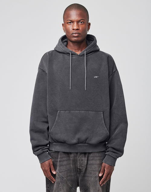 A person with short hair and a neutral expression stands facing forward in a dark gray LIVE FAST DIE YOUNG LFDY Eagle Hooded sweatshirt (hood down, oversize fit) and matching jeans. The background is plain light gray.