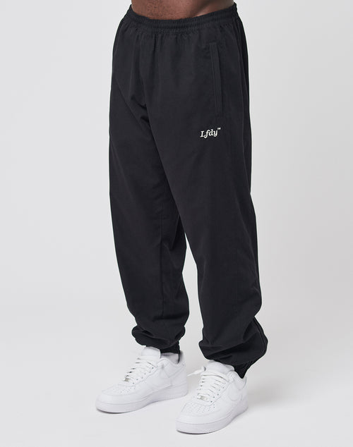A person wearing black LFDY Trackpants by LIVE FAST DIE YOUNG, featuring white LFTDY embroidery on the left thigh and made from a comfortable polyester blend, pairs them with white sneakers against a plain white background.